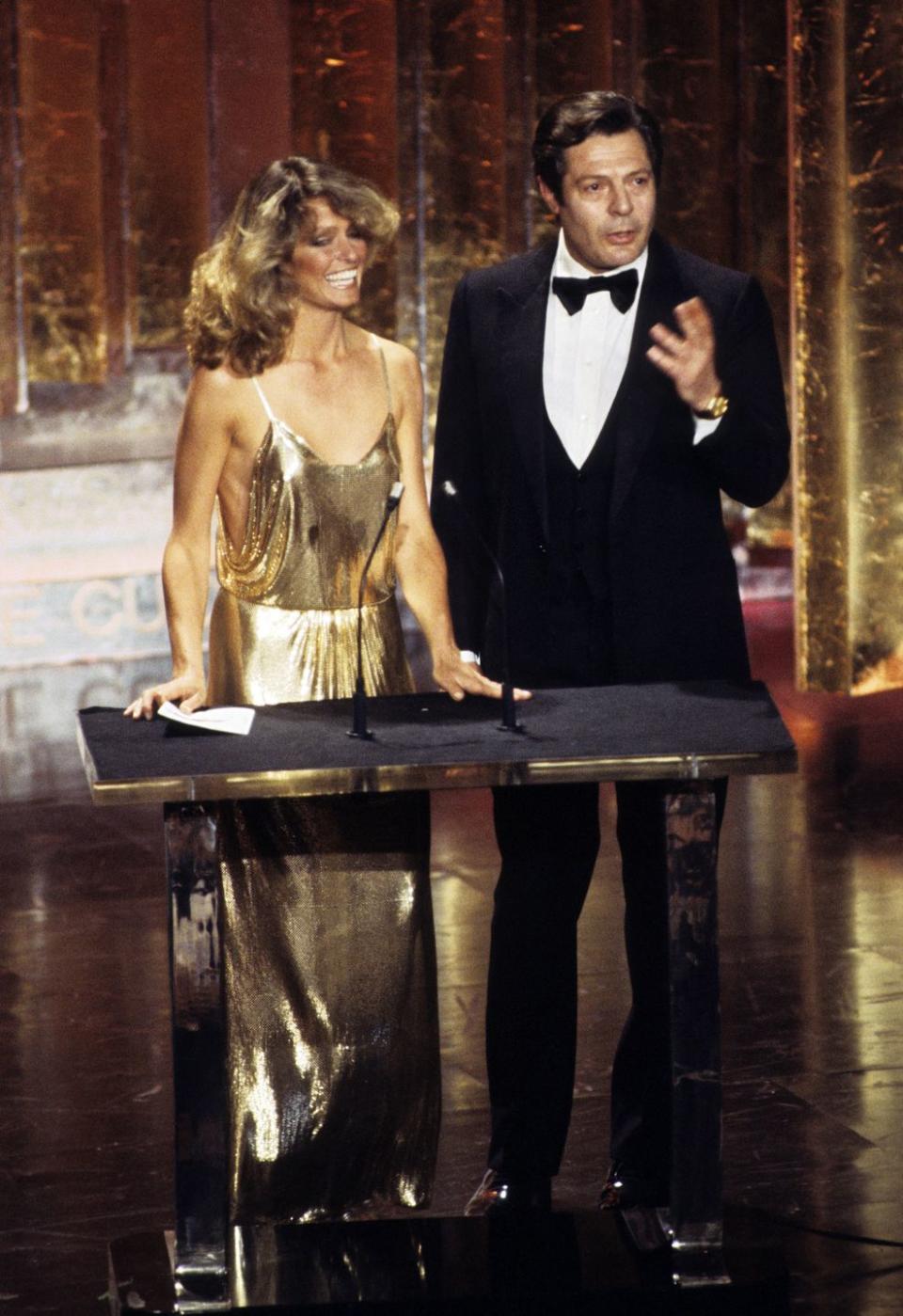 <p>Farrah Fawcett wore a metallic gold Halston dress to the 50th Annual Academy Awards. The <em>Charlie’s Angels</em> actress is shown here with Italian actor Marcello Mastroianni.</p>