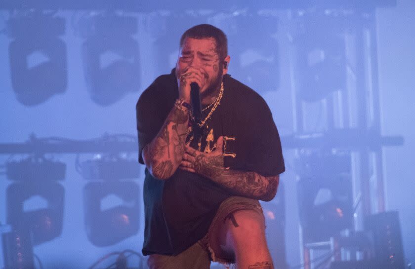 A man with tattoos on his arms and face crouches while singing onstage