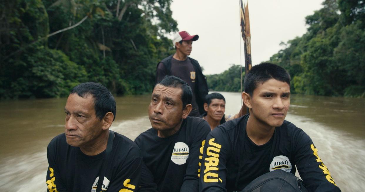 Five Indigenous men sit in a boat racing toward the camera, all looking slightly away