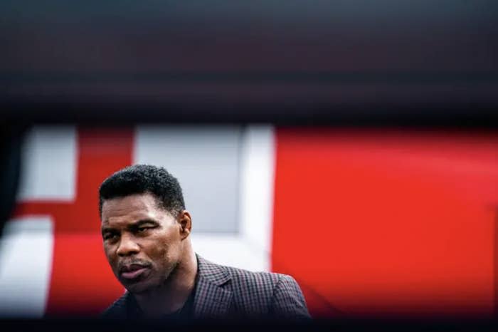 Herschel Walker in a gingham suit against a red background; the top part of the shot is covered by a dark wall that's out of focus