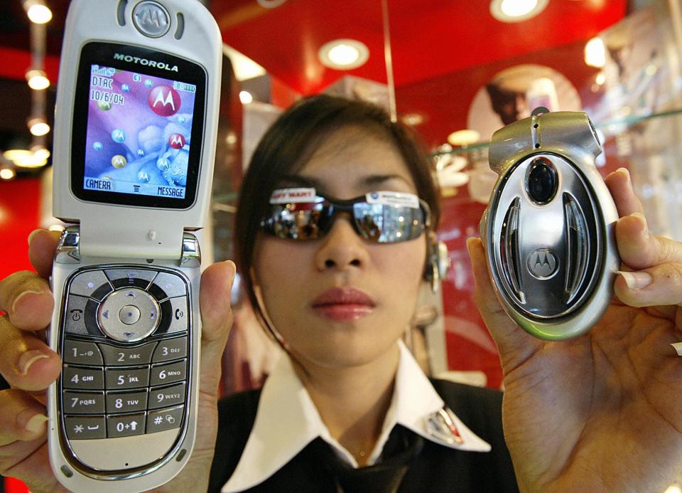 a woman with silver sunglasses holds up the Motorola V600 flip phone in one hand, and a silver Motorola Bluetooth speakerphone in her other hand