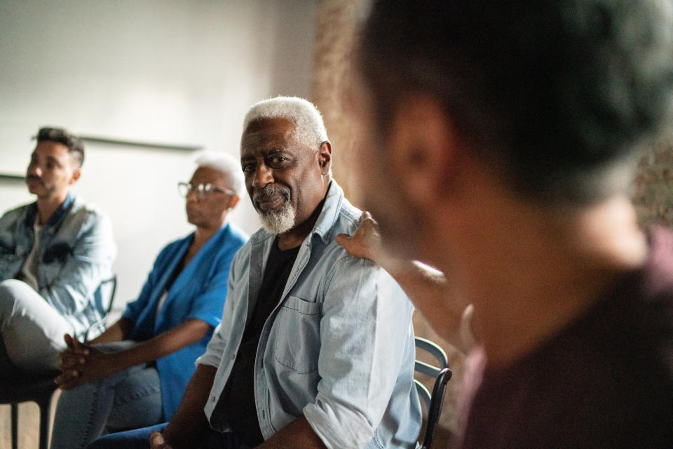 A group of people sit in a circle in a meeting. One person places a hand on the shoulder of another, offering support