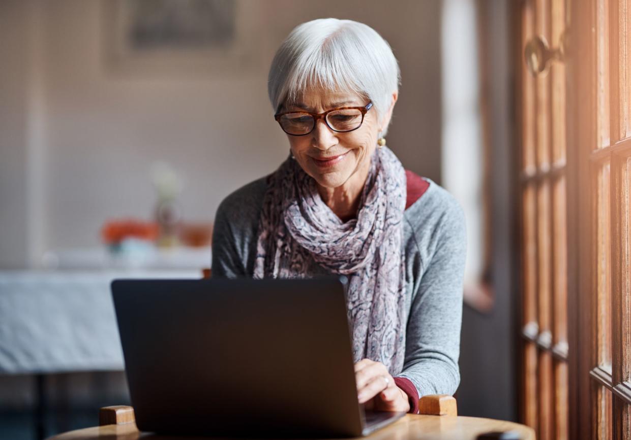 Senior woman on laptop at a small round table in a retirement home with large window doors to her right, with a blurred background of a dimly lit living area