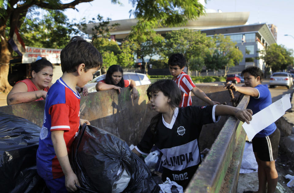 In this June 26, 2012 photo, members of two families, from left, Perla Soria, Romel Matechip, Maria Belen Britez, Victoria Nunez, Roger Britez, and Sara Aquino search in a trash container for things to recycle, as well as food, along the "15 de Agosto" street next to the Legislative Palace, behind, in Asuncion, Paraguay. Across the street from the gleaming Legislative Palace lies a trash-strewn tent camp of Paraguay's landless poor, where pigs root in the dirt and flames lap at the grate of a rudimentary cooking pit. The stark contrast in the capital tells the story of a divided people living under the same red-white-and-blue-striped flag. (AP Photo/Jorge Saenz)