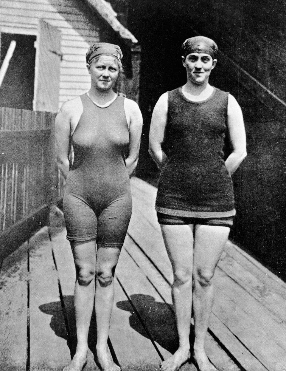 Two female swimmers standing on a wooden patio pose for a black and white photo in front of a house