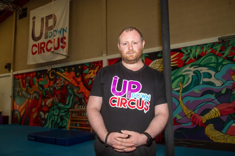 Chris Moore leans on a pillar with his hands clasped together. He has a short beard and is wearing a T-shirt with the Up Side Down Circus logo in multi-coloured lettering