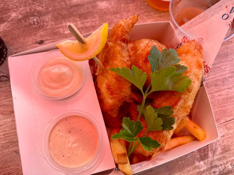 A box of fried fish and fries with a wedge of lemon and some garnish, accompanied by two sauces.