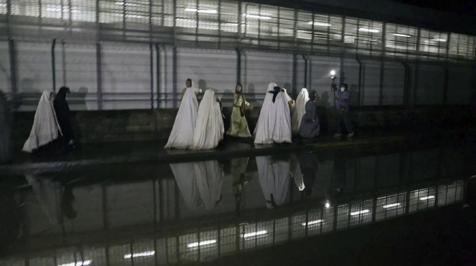 A group people, some cloaked from head to toe in white robes, walk on a sidewalk near a wall with lighting