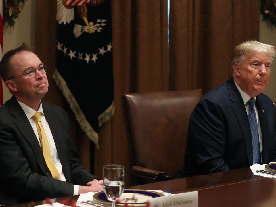 Mick Mulvaney and Donald Trump seated in front of flags