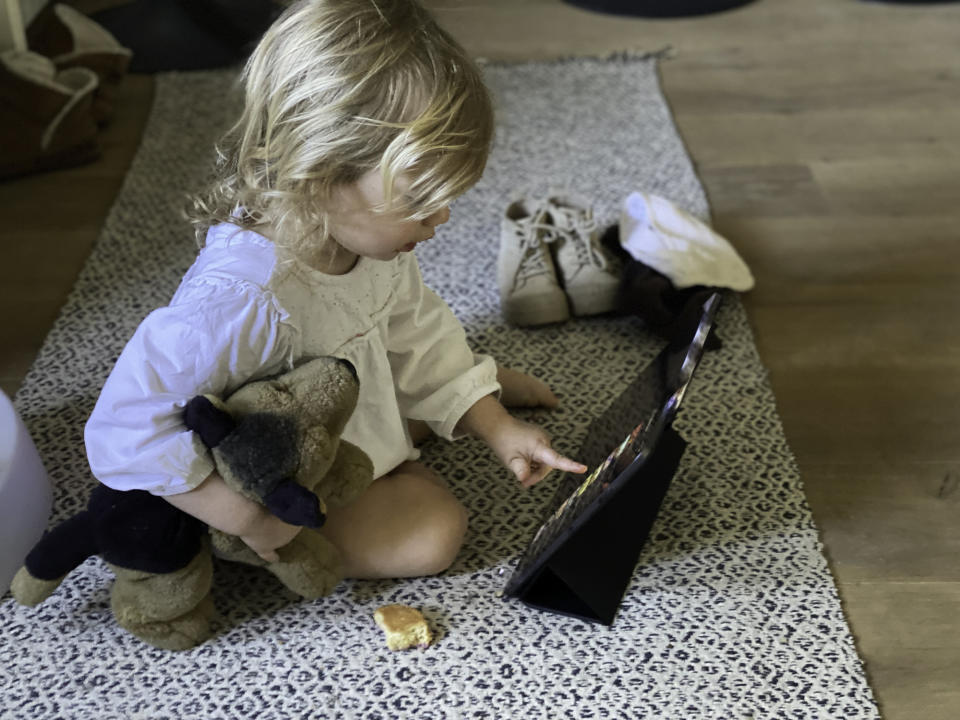 A little girl holding a stuffed teddy plays on an iPad.