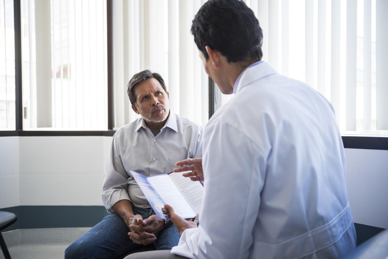 A white male doctor presumably asking his seated bearded, white male patient about access to guns. 