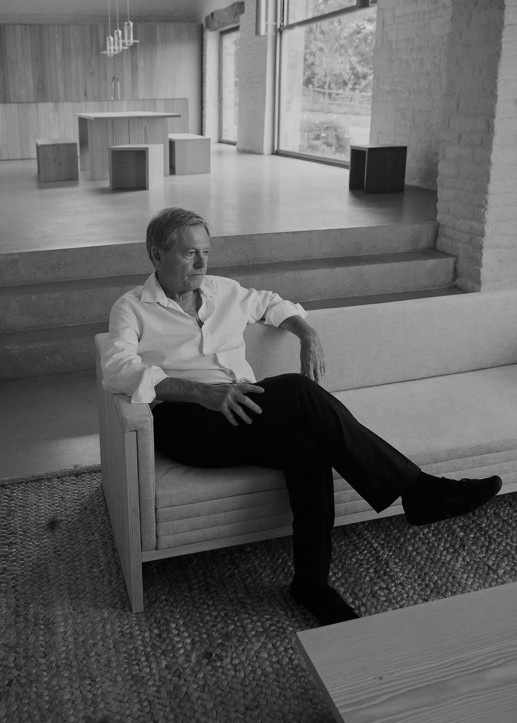 a man in a white shirt and dark pants sitting on a wood frame daybed with spare furnishings around him