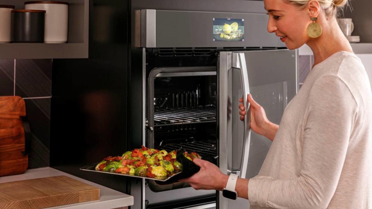  A woman holding a tray of vegetables and opening the door of a GE Appliances wall oven. 