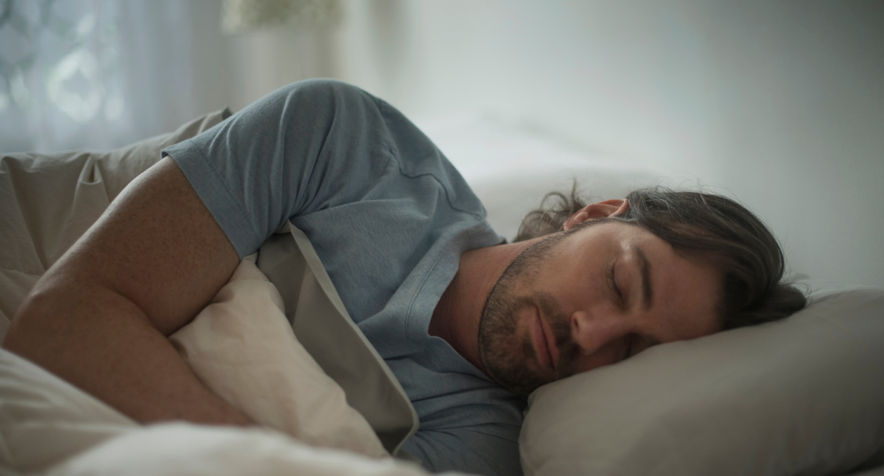 man with beard and brown hair wearing blue t-shirt sleeping in bed, pillow and sheets, sleeping on his side