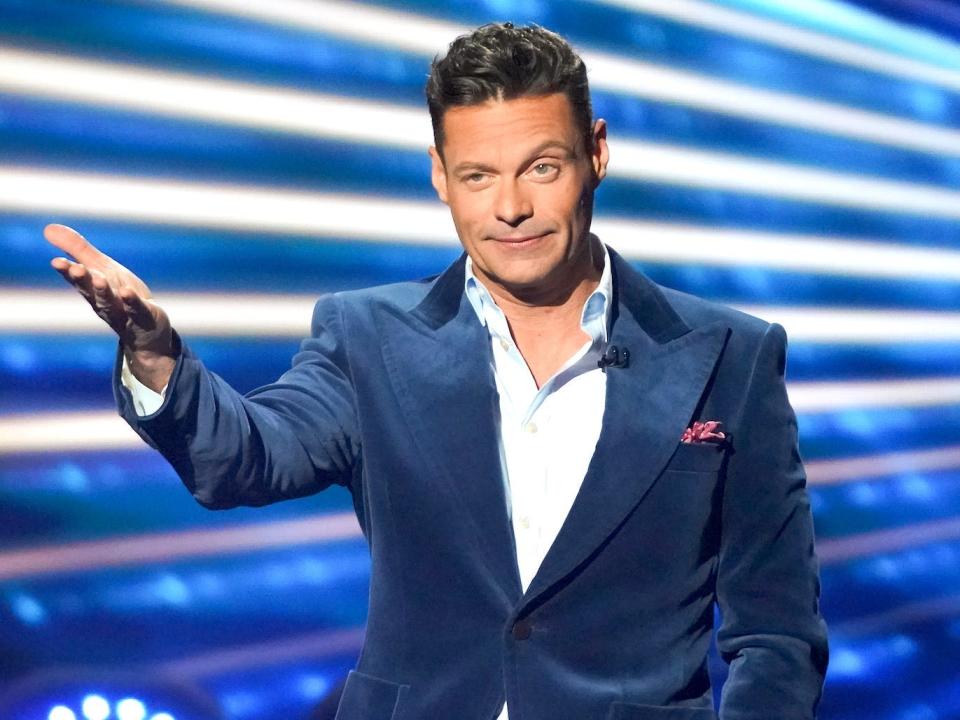 A man (Ryan Seacrest) wearing a blue velvet blazer and grey pants on the stage of "American Idol."