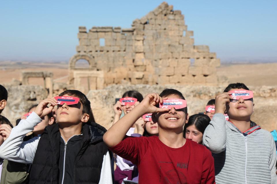 Bunch of people look up towards the sun while holding pink eclipse glasses up to their eyes in front of a sand colored brick wall in a desert landscape