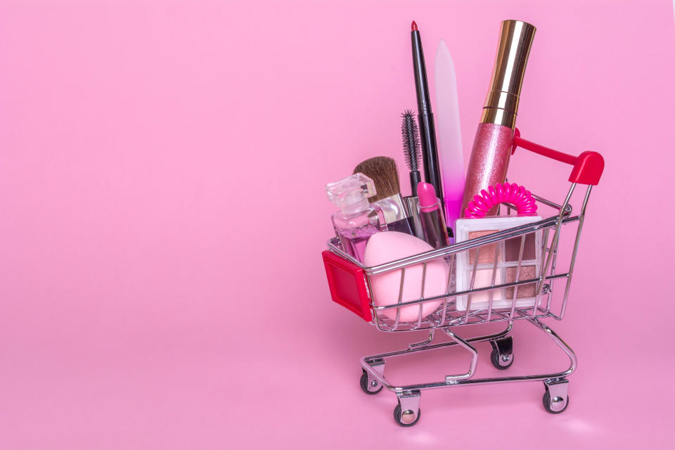 Shopping trolley with makeup on a pink background. Perfume, sponge, brush, mascara, pencil, nail file, eye shadow, lip gloss in the basket