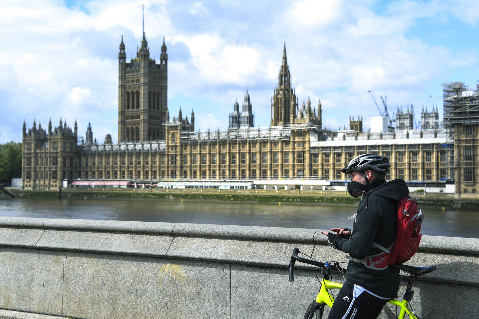 FILE - In this file photo dated Friday, May 1, 2020, a man stands with his bike, wearing a protective mask to protect against coronavirus, on the south bank of the River Thames, against the backdrop of the Houses of Parliament, in London, as the country continues its lockdown to curb the spread of coronavirus. The government has decided to scrap a remote-voting system used during the coronavirus pandemic, and has summoned lawmakers back to parliament on Tuesday June 2, 2020, but many aren’t happy with the arrangements. (AP Photo/Alberto Pezzali, FILE)