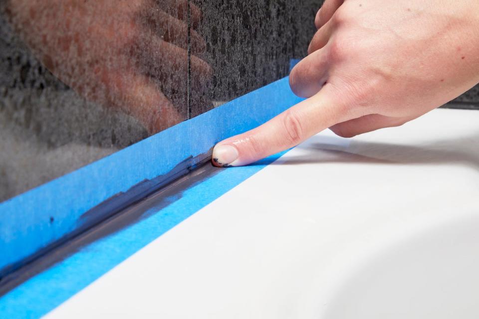 Woman uses her finger to smooth the bead of caulk; painter's tape keeps it off the tile.