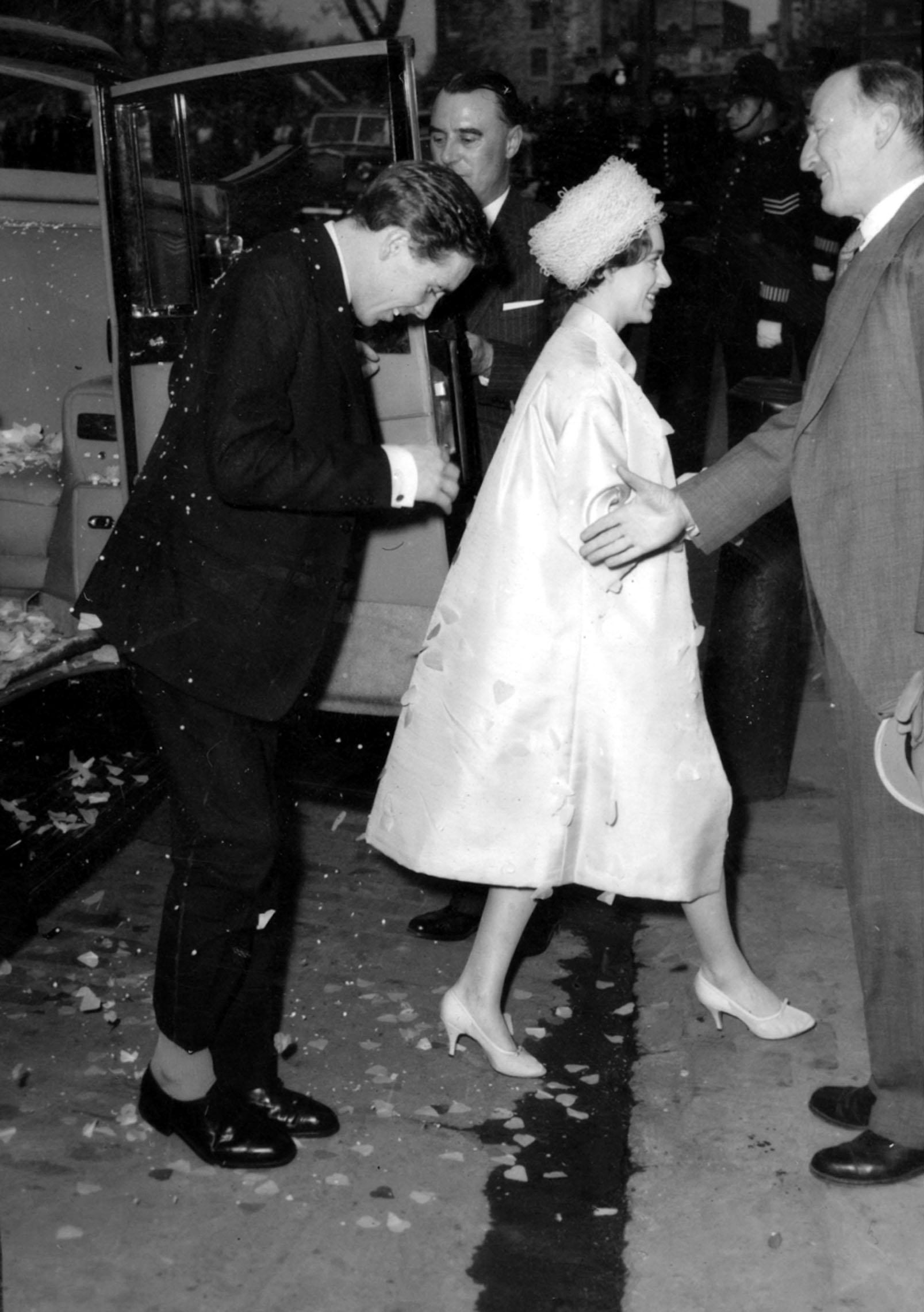 Antony Armstrong-Jones brushes vigorously at the confetti and rose petals which cling to him as he and his bride, Princess Margaret, are welcomed at Tower Pier by Viscount Simon, Chairman of the Port of London Authority, prior to the embarkation in the Royal Yacht Britannia and their West Indies honeymoon.