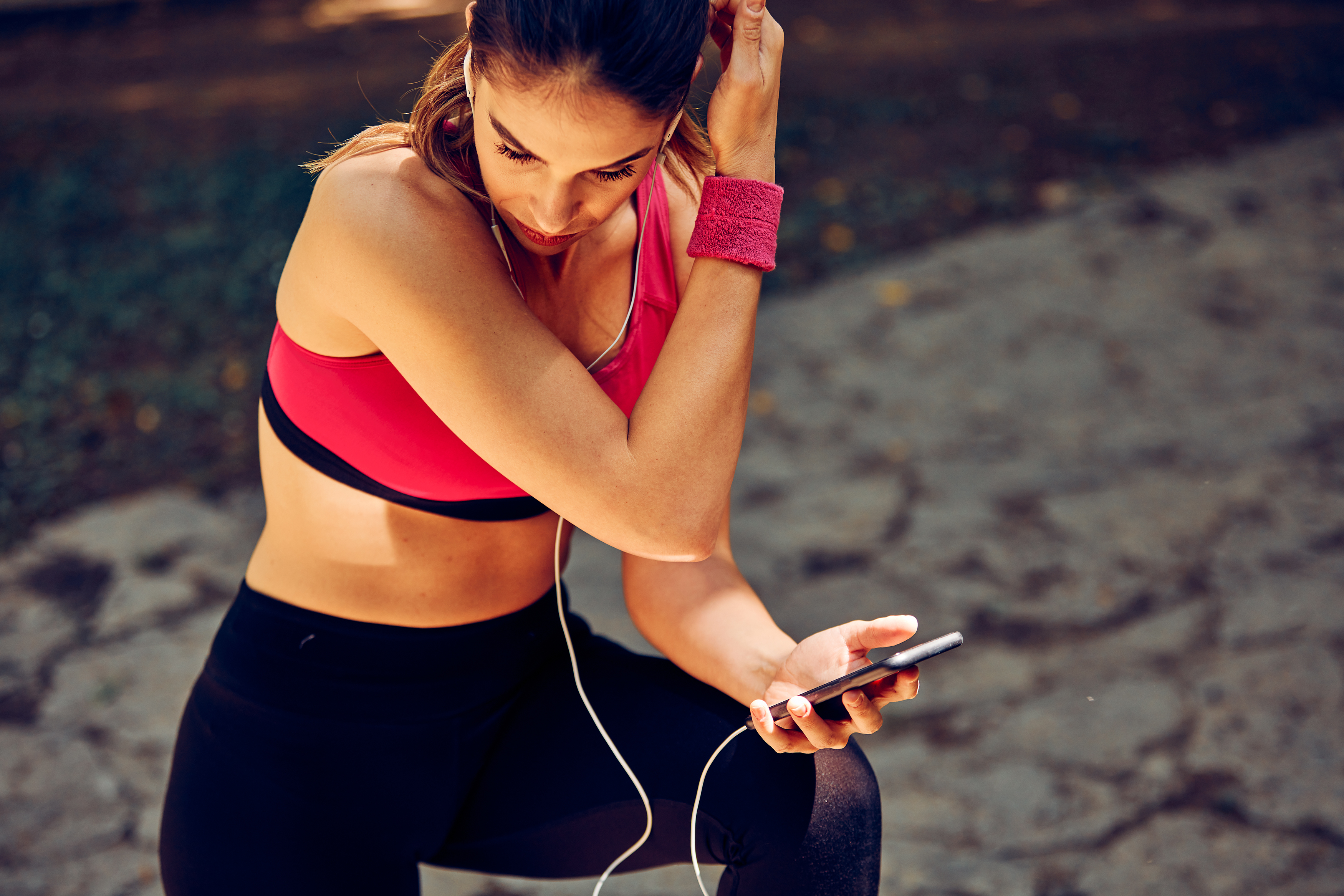 Smiling sportswoman crouching, taking a break, and using phone after running in nature." data-uuid="198814c9-3fe6-3618-8d5d-4c7d4cb58a34