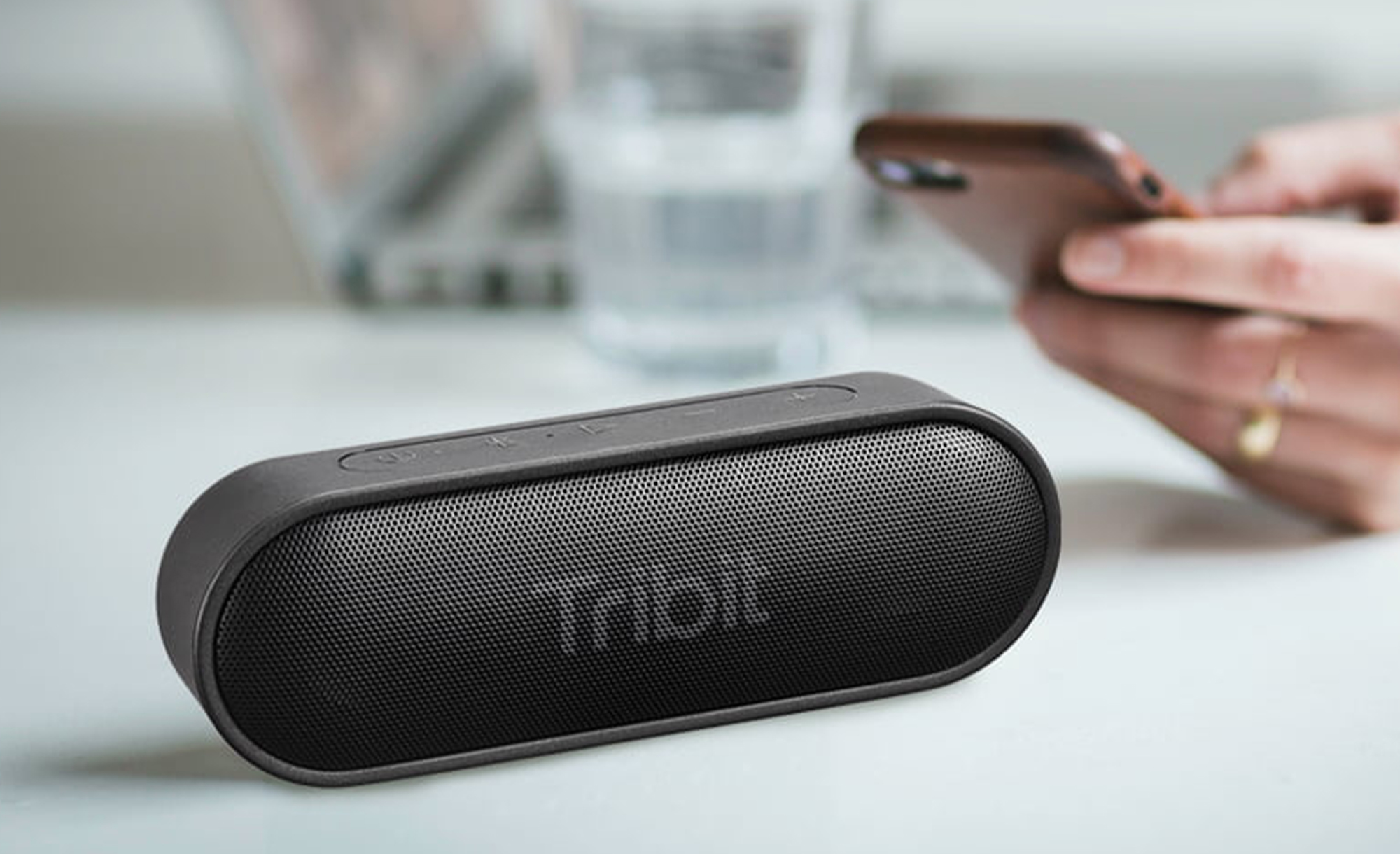 A Tribit XSound Go speaker sitting on a white table with a person using a smartphone in the background.