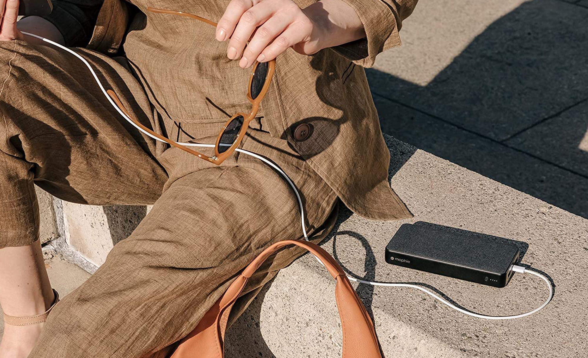 A person sits on concrete steps in the sunshine with a Mophie Powerstation XXL charging their phone next to them.