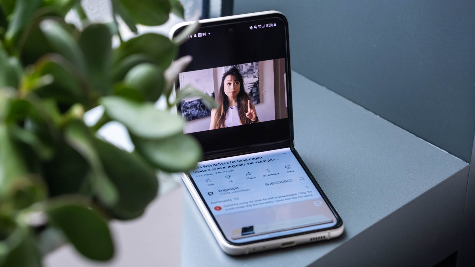 The Samsung Galaxy Z Flip 3 folded to prop itself up on a table, with the top half of its screen showing a YouTube video and the bottom showing the title, likes, dislikes, channel, top comment and suggested videos.