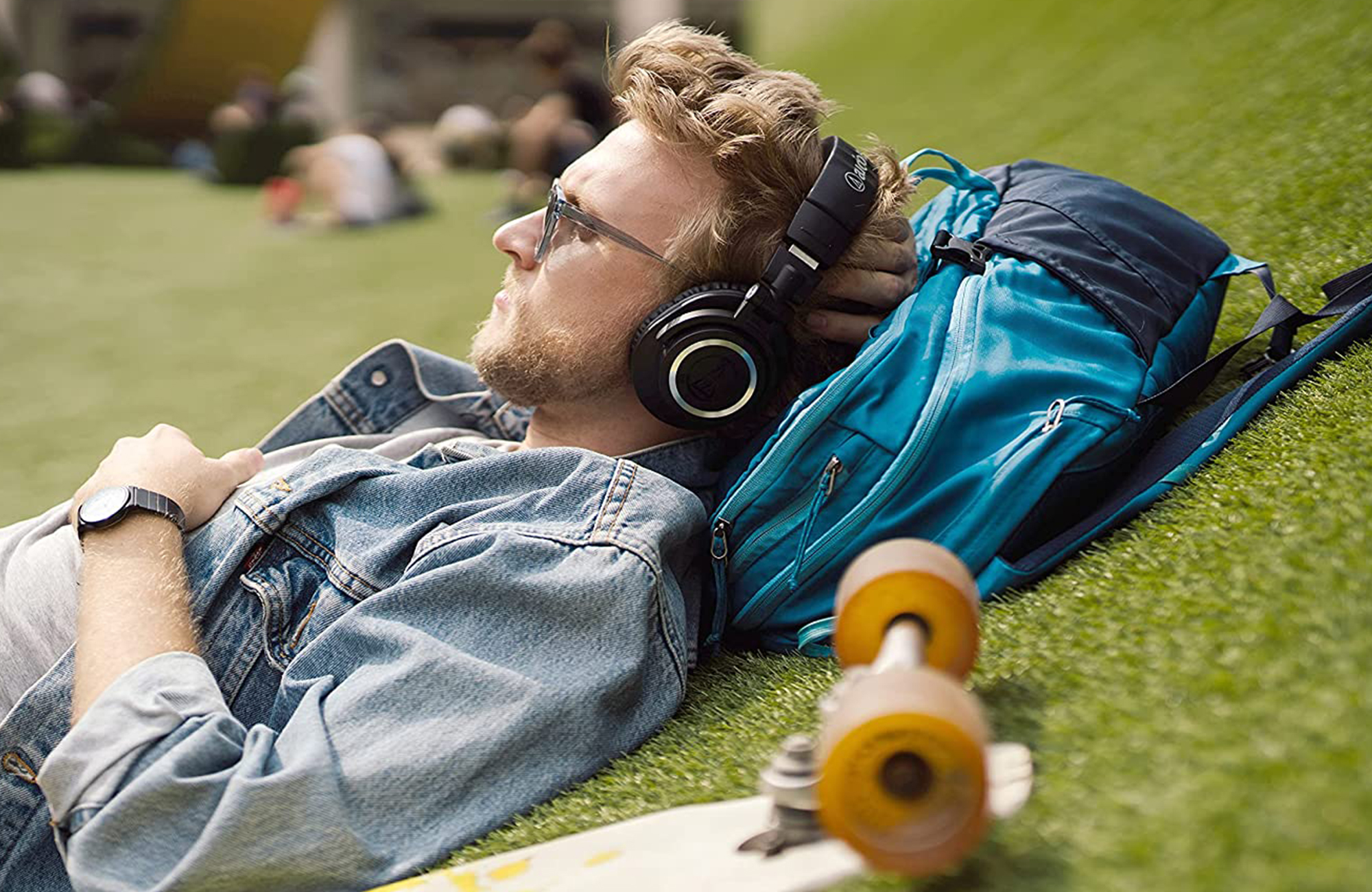 Audio-Technica ATH-M50xBT2 for the Engadget 2021 Holiday Gift Guide. A man wearing the headphones lies on a grassy hill using a backpack for a pillow. 