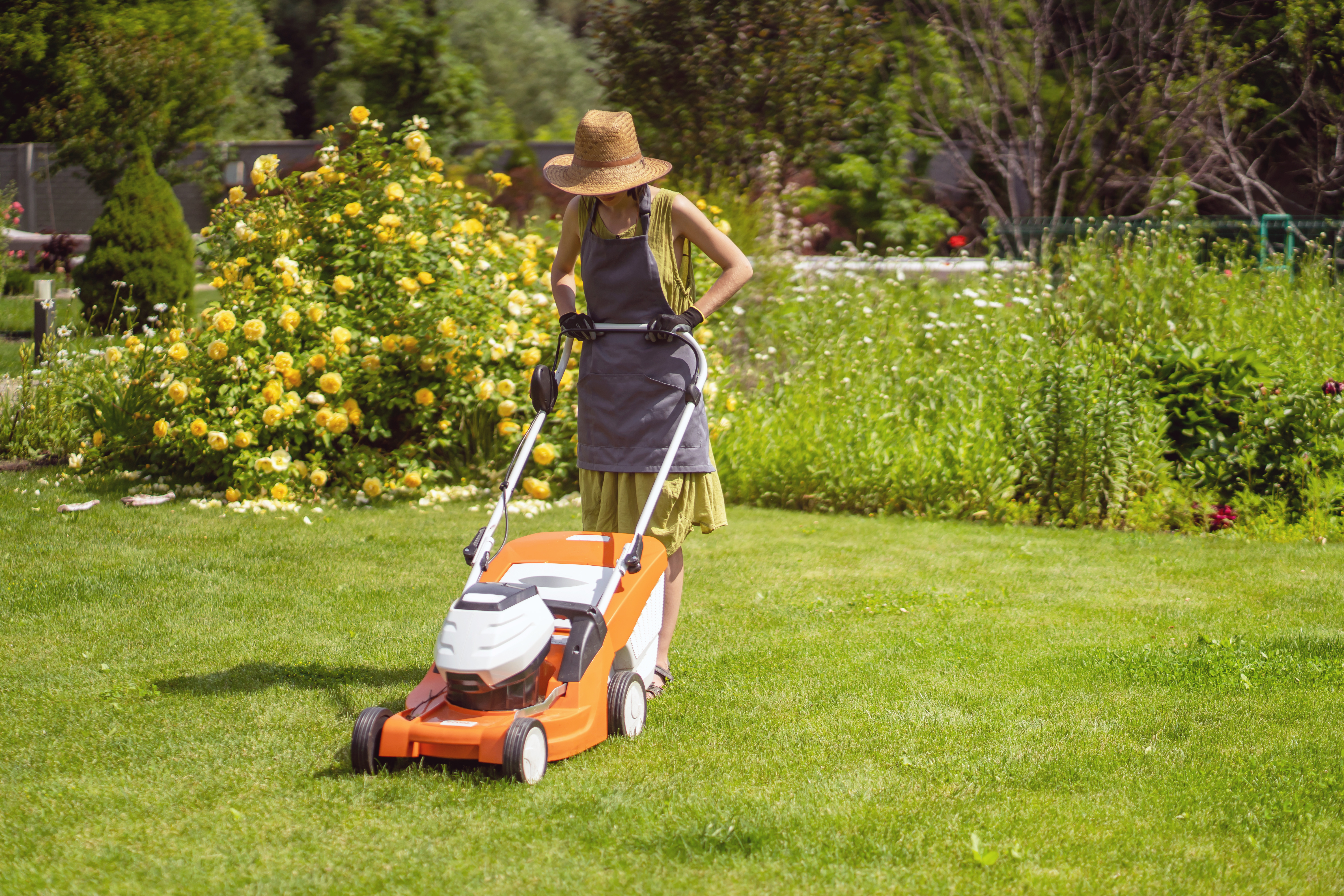 A young girl in a straw hat is mowing a lawn in the backyard with an orange lawn mower. A woman gardener is trimming grass with the grass cutter. A lawnmower is cutting a lawn on a summer sunny day.