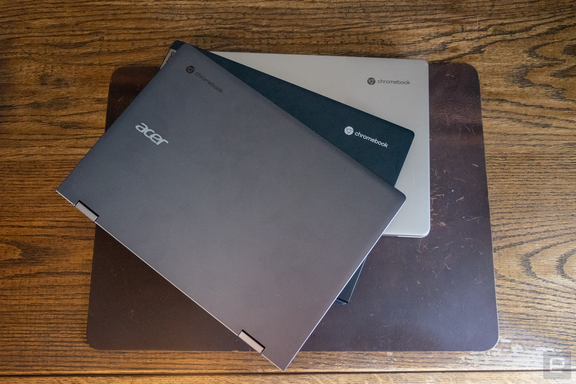 Three Chromebook laptops stacked on top of each other, fanning out a bit, on top of a desk protector on a wooden table." data-uuid="24972f7b-1262-3d93-8e2b-f189a92fda1b