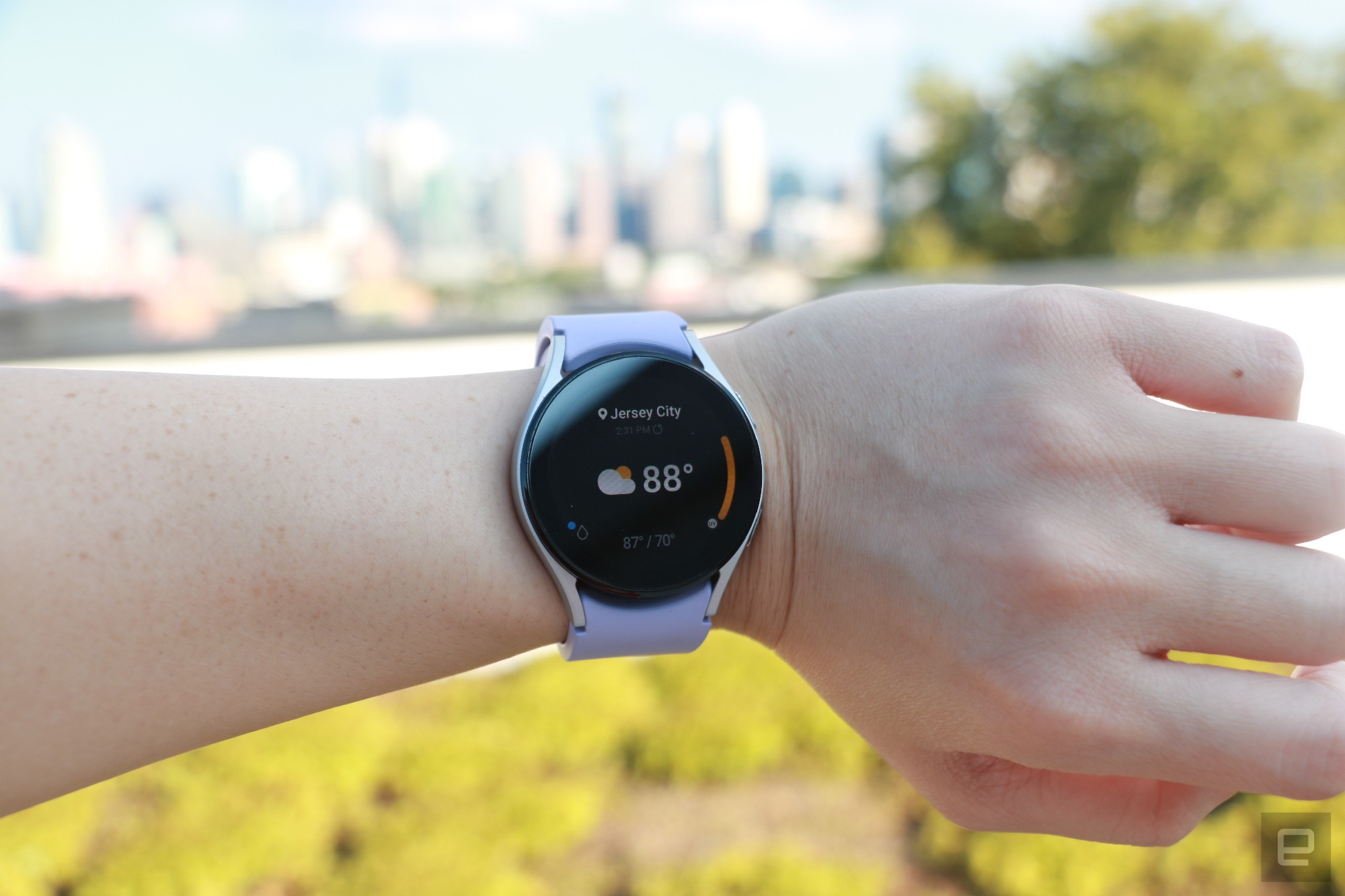 The Samsung Galaxy Watch 5 on a wrist, showing the weather app. It says Jersey City at the top, followed by 88 degrees in bigger font taking up the majority of the page, and a cloud with sun icon on its left.