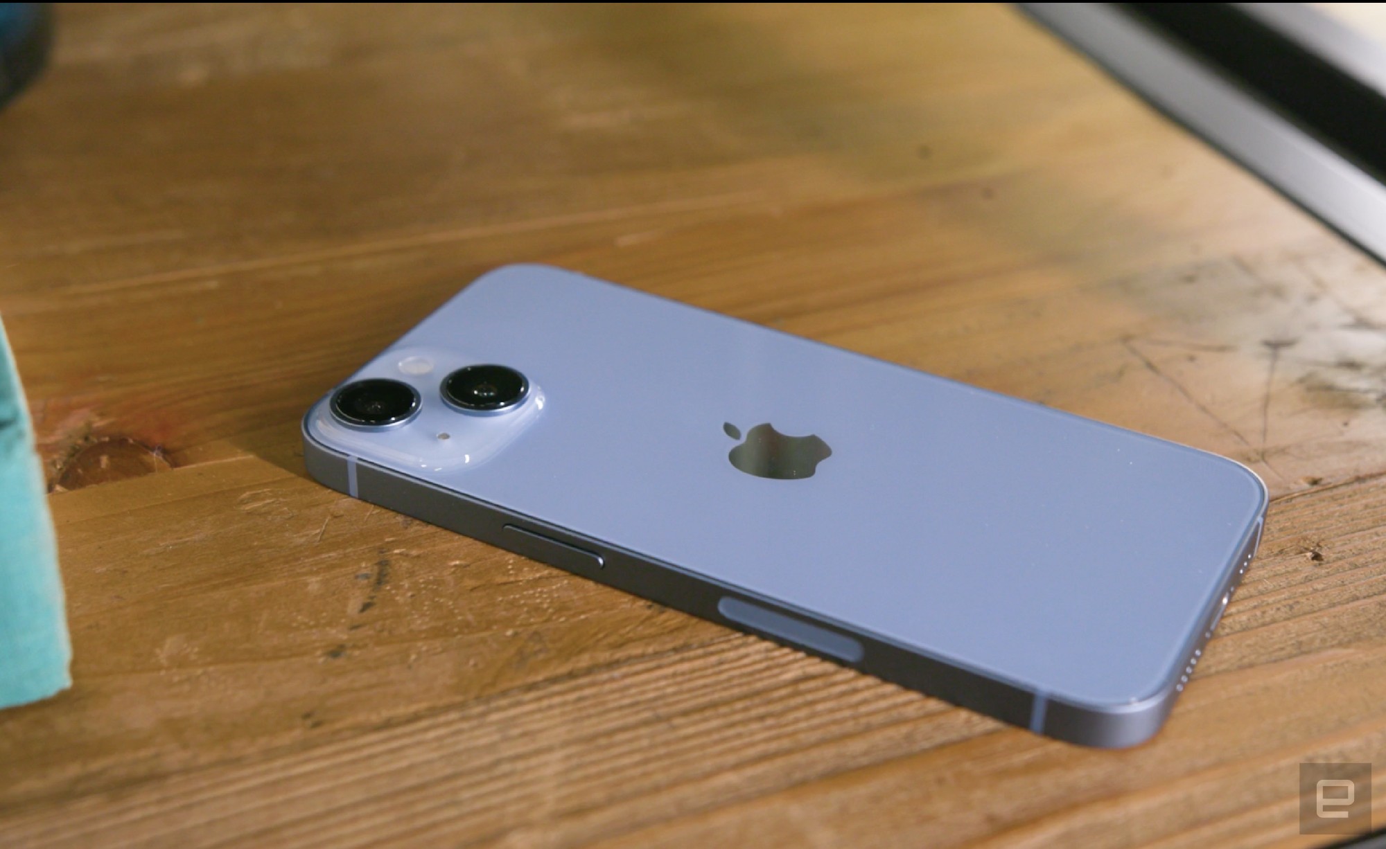The blue iPhone 14 laying face down on a wooden surface, with its right edge facing the camera.