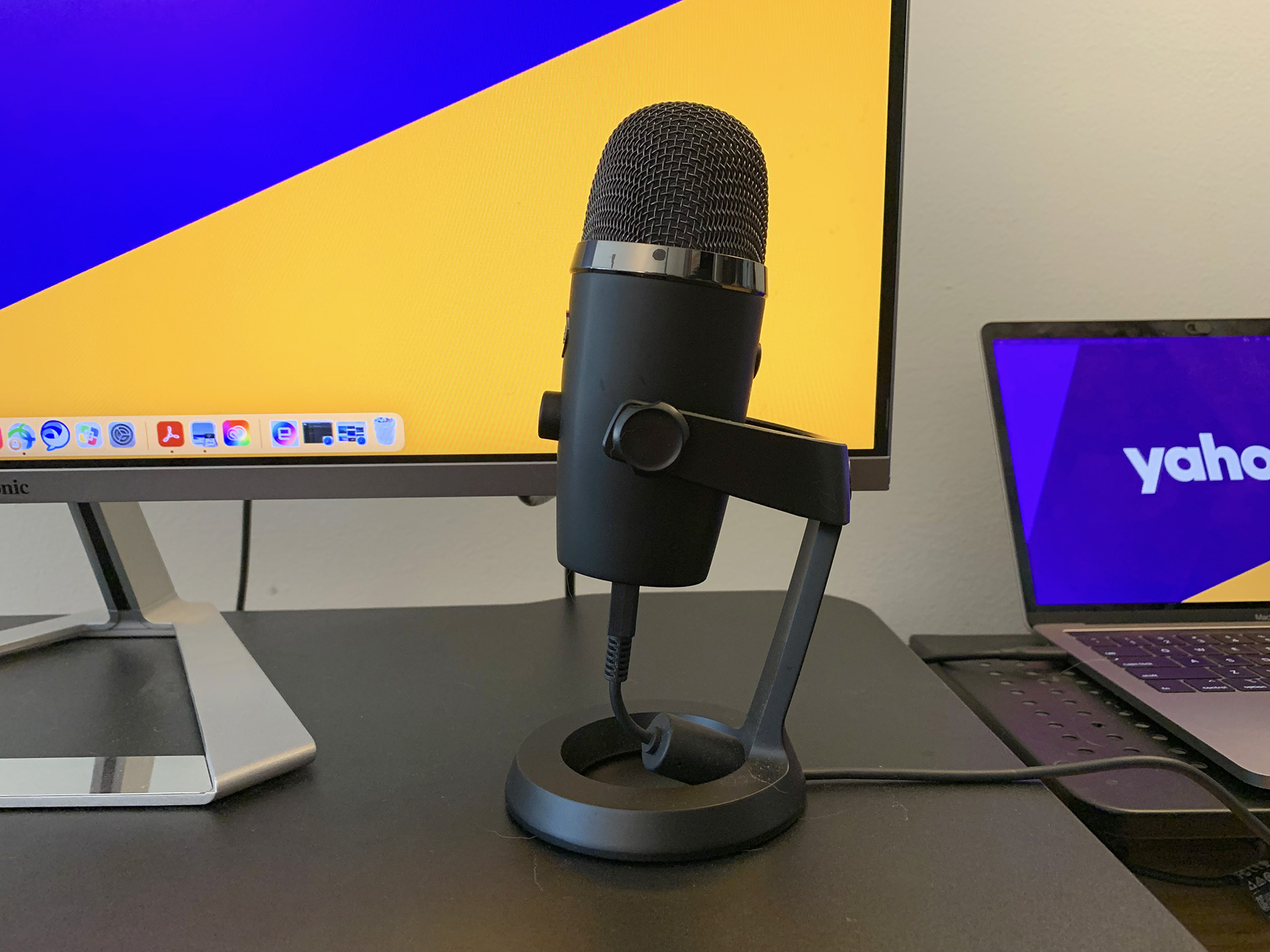 The Blue Yeti Nano microphone on a black desk riser in front of a laptop and a computer monitor.