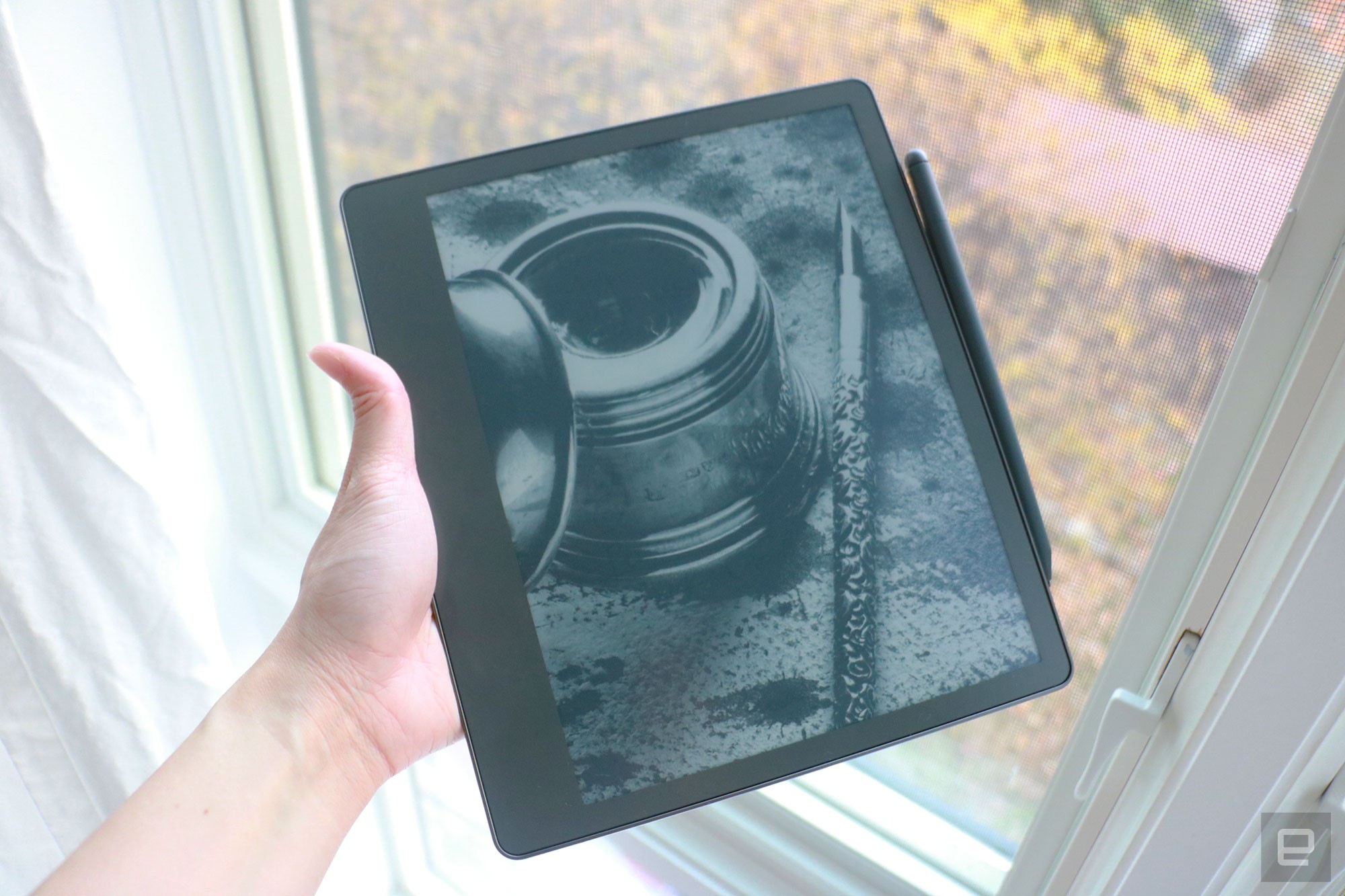 The Amazon Kindle Scribe held in mid-air by a hand gripping its left side, with a pen attached to the right side. In the background is a window with white curtains.