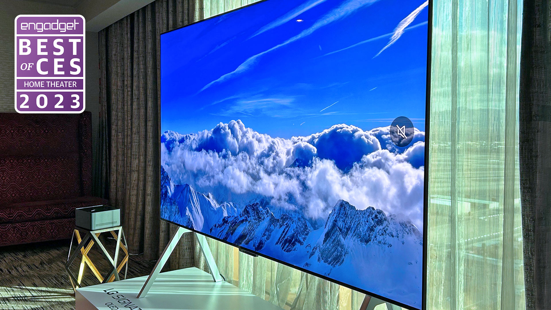 An angled view of the 97-inch LG M3 OLED with wireless tech sitting in a hotel room in front of a translucent window curtain." data-uuid="19e83474-b8ff-30b7-b8f9-c6b5a0606b8e