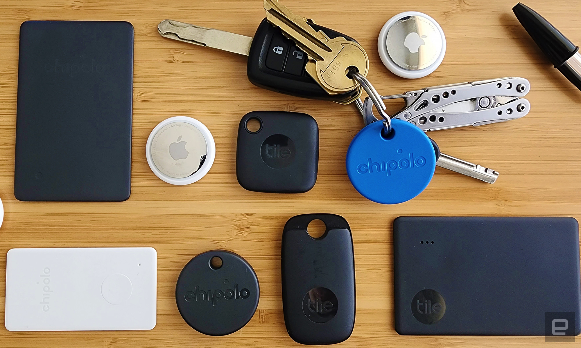 An assortment of bluetooth trackers arranged in a grid on a wooden background. Trackers include black Tile trackers in various shapes, two silver and white AirTag trackers and a round blue Chipolo tracker attached to a set of keys with a multitool key chain. " data-uuid="e92fd876-b075-3bd1-9c92-355d3b241508