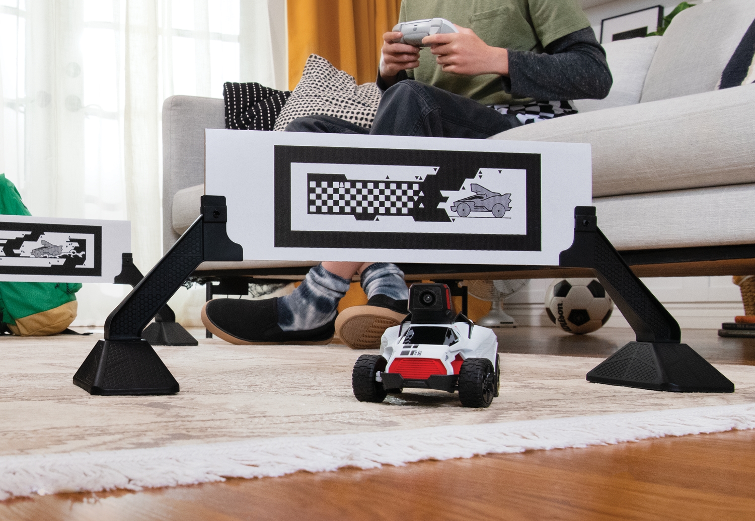 A toy RC car driving under a “gate” in a person’s living room. A child is holding a PlayStation controller while sitting on a couch behind it.