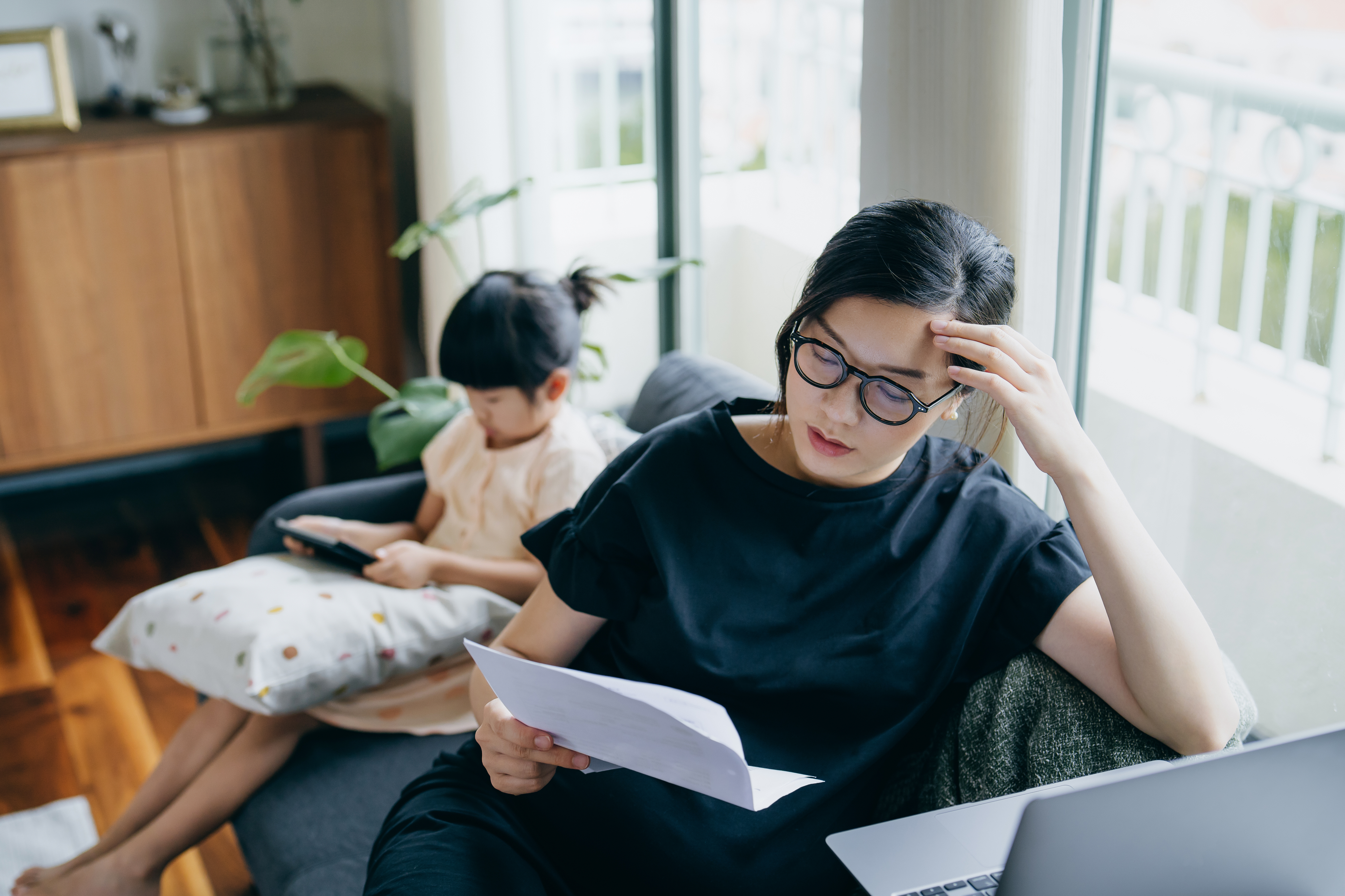 Young Asian mother looking stressed while managing her financial bills and tax documents, working from home on laptop and her daughter is using digital tablet in the background. Working mother managing work life and childcare at home