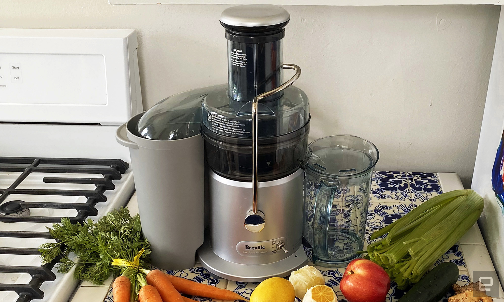 The Juice Fountain Plus juicer sits on a blue and white countertop surrounded by celery, carrots, apples, lemons and cucumbers. 