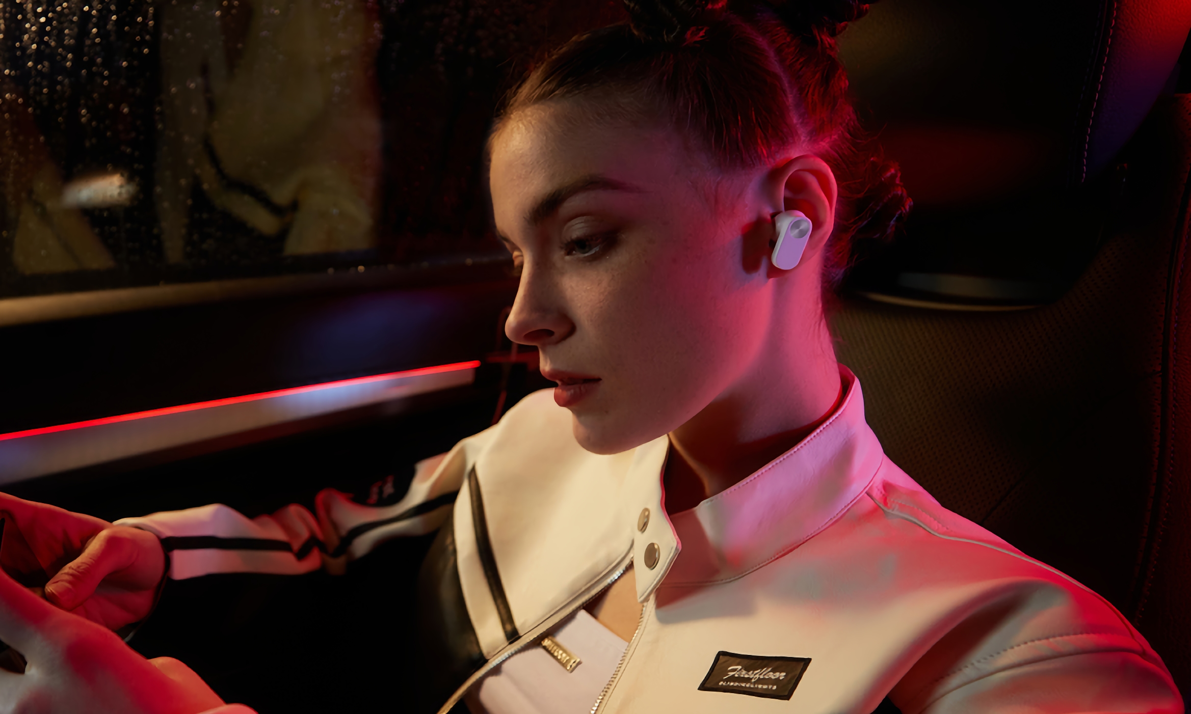 A woman riding as a passenger in a car at nighttime, wearing white Nord Buds 2 earbuds.