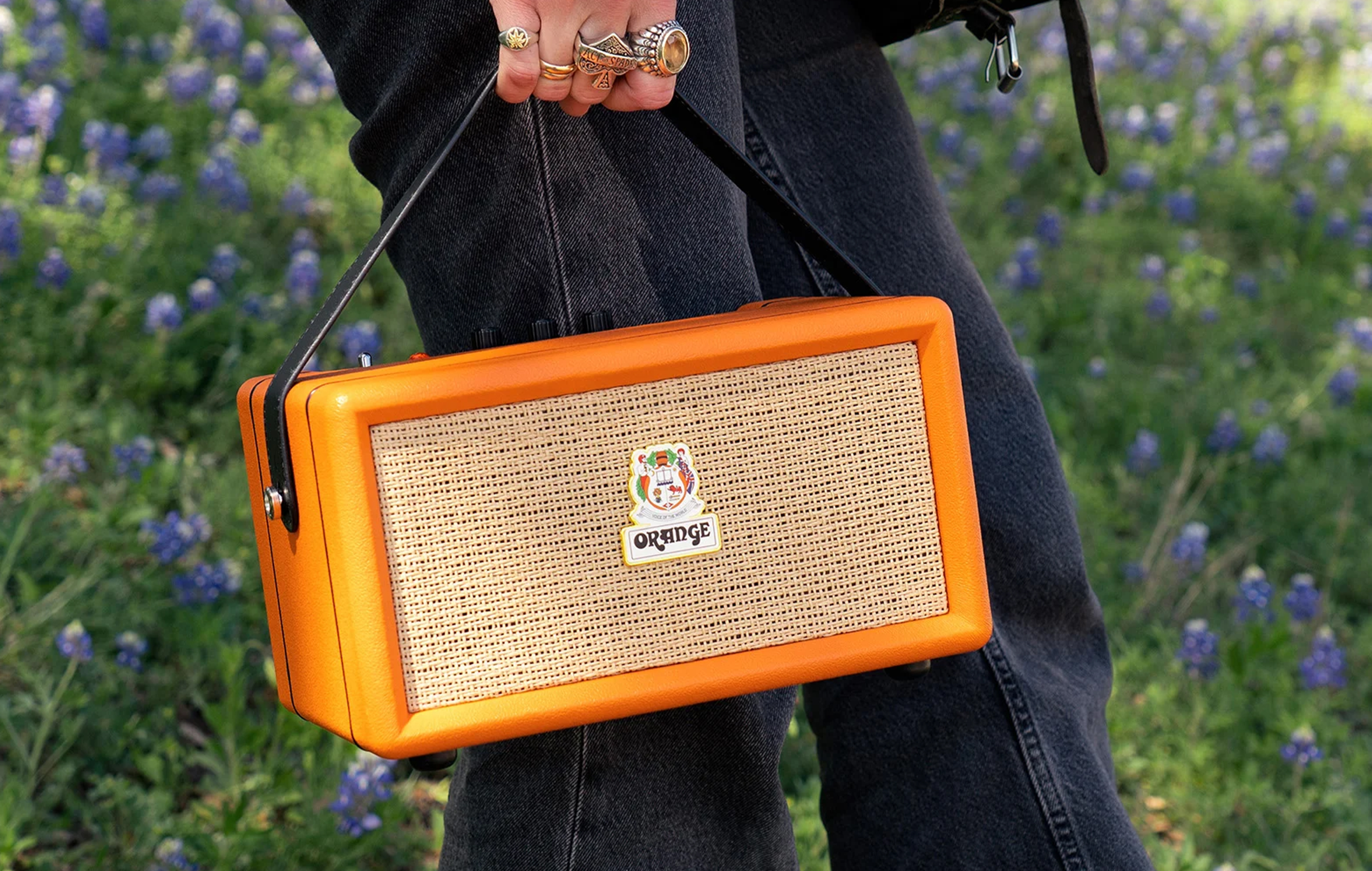 A cropped image shows someone in jeans and wearing chunky rings holding the Orange Amps Box portable Bluetooth speaker by its carrying strap.