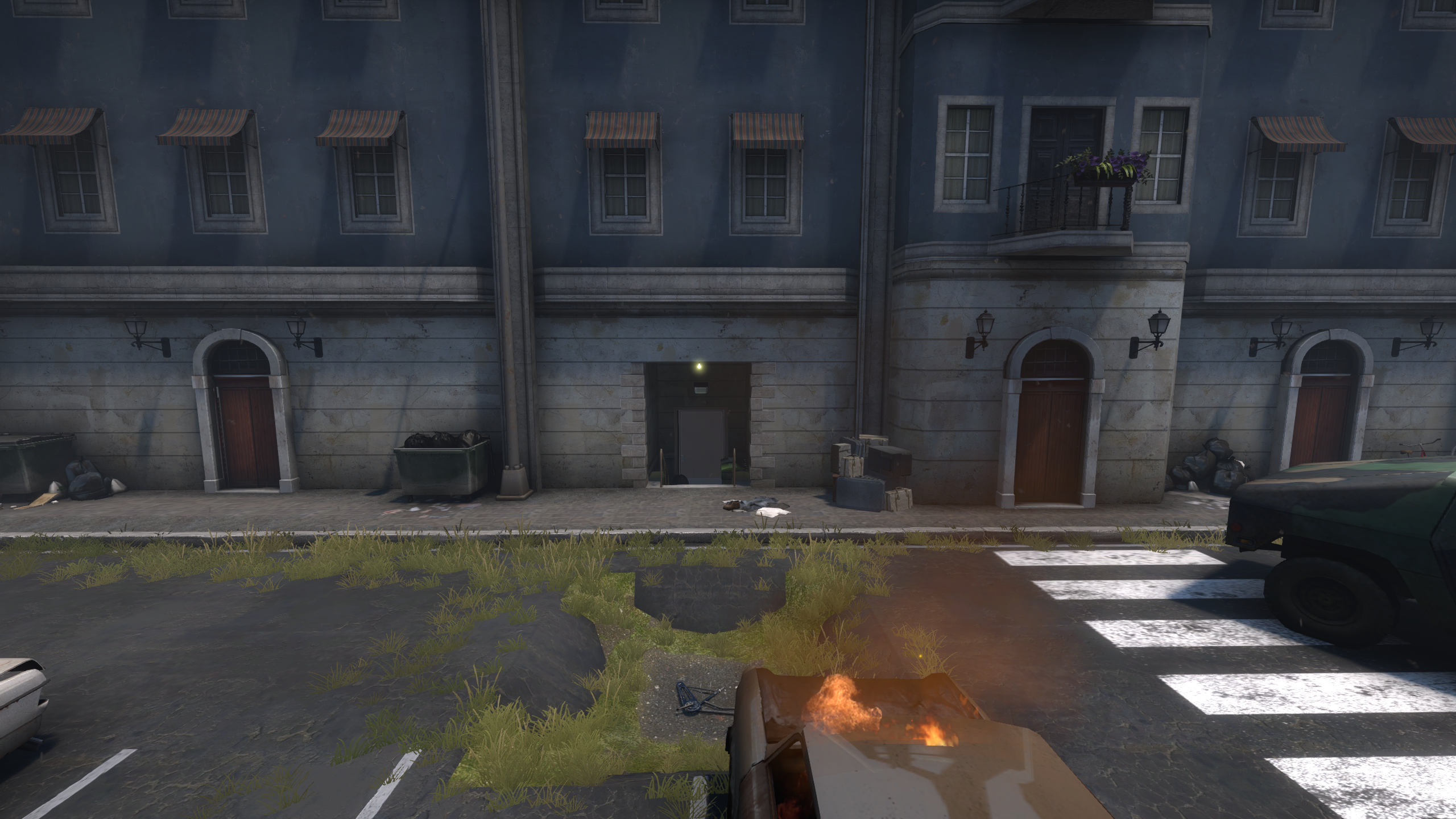 Video game screenshot from ‘Counter-Strike: Global Offensive.’ On the side of a war-torn building is a dark passageway with a light over its entrance. A flaming car sits in the foreground.