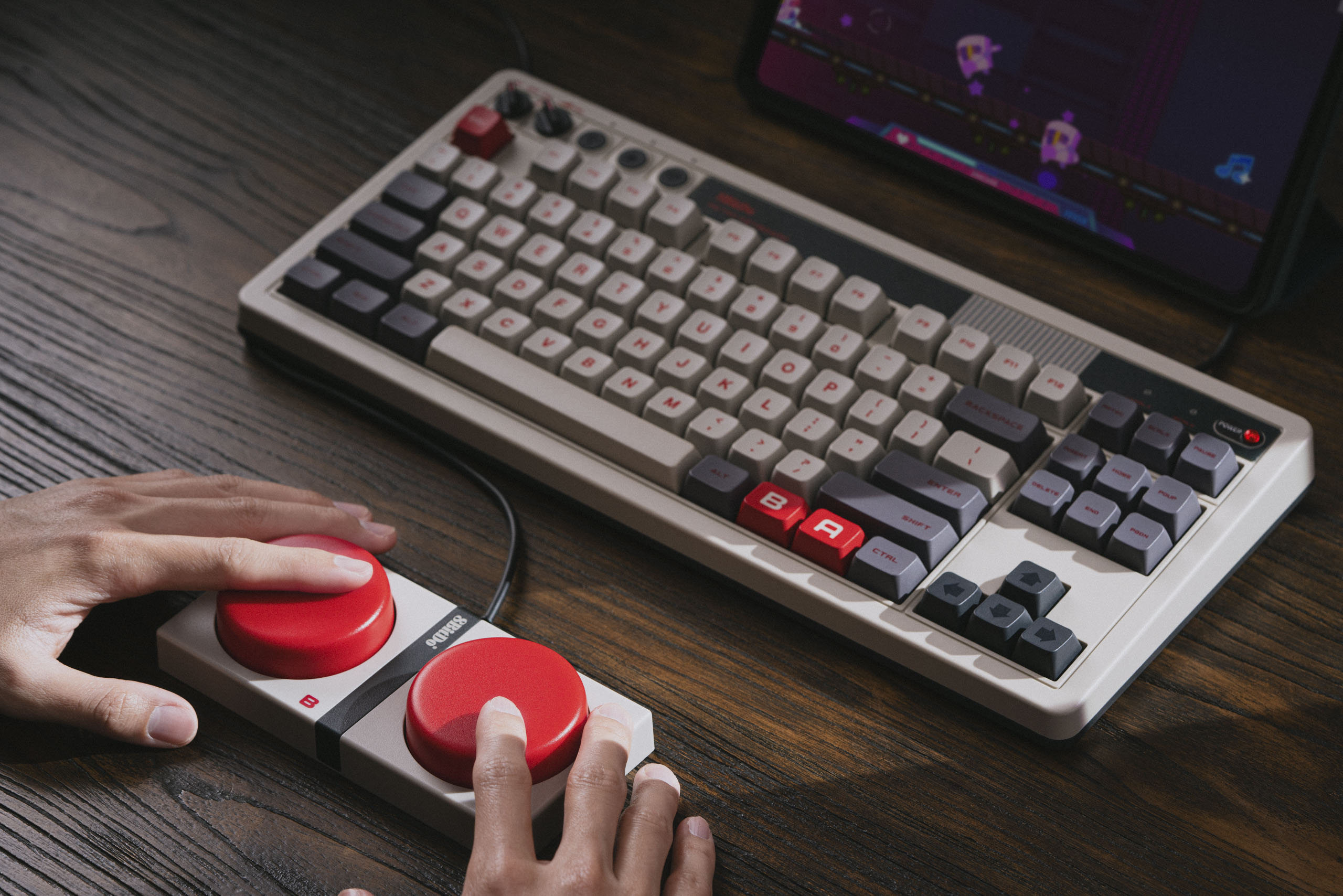 Marketing photography. A NES-colored mechanical keyboard sits on a dark wooden desk. Two hands mash two giant red buttons (also NES-styled) below.