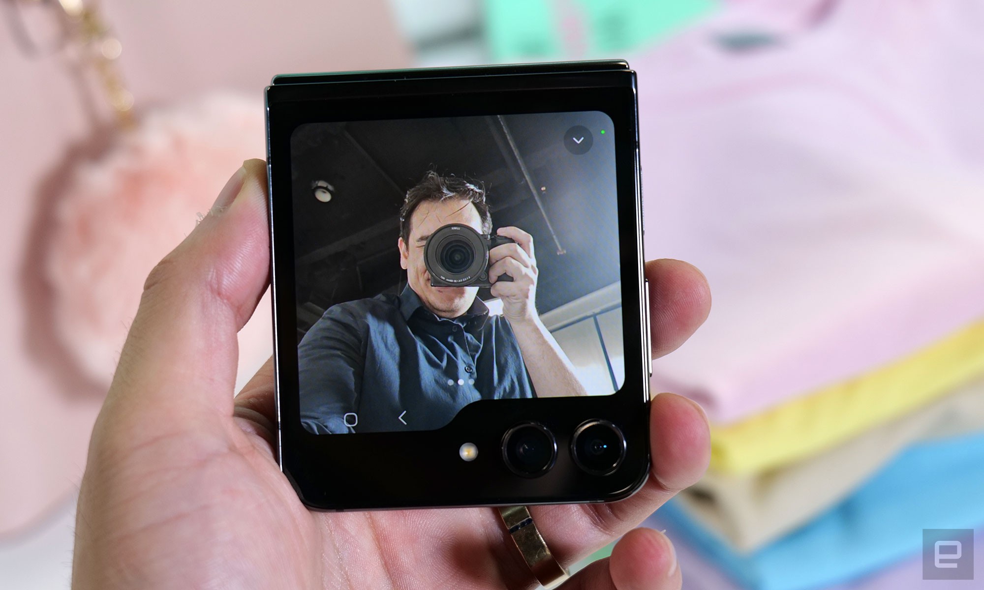 The Samsung Galaxy Z Flip 5 folded in half, with the camera preview appearing on its cover screen facing the camera. In the viewfinder is a man using a DSLR to take a photo of the phone.