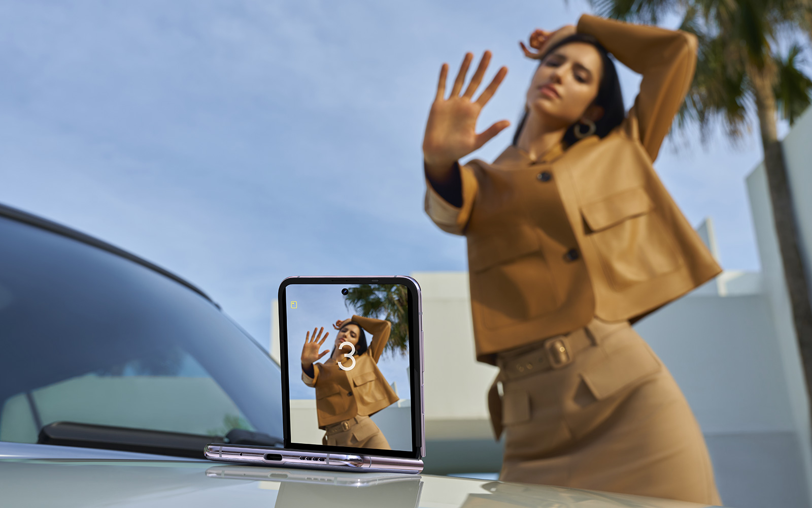 A half-opened Tecno Phantom V Flip phone placed on a car hood, with its main camera facing a model who is posing with a hand gesture to remotely trigger the shot.