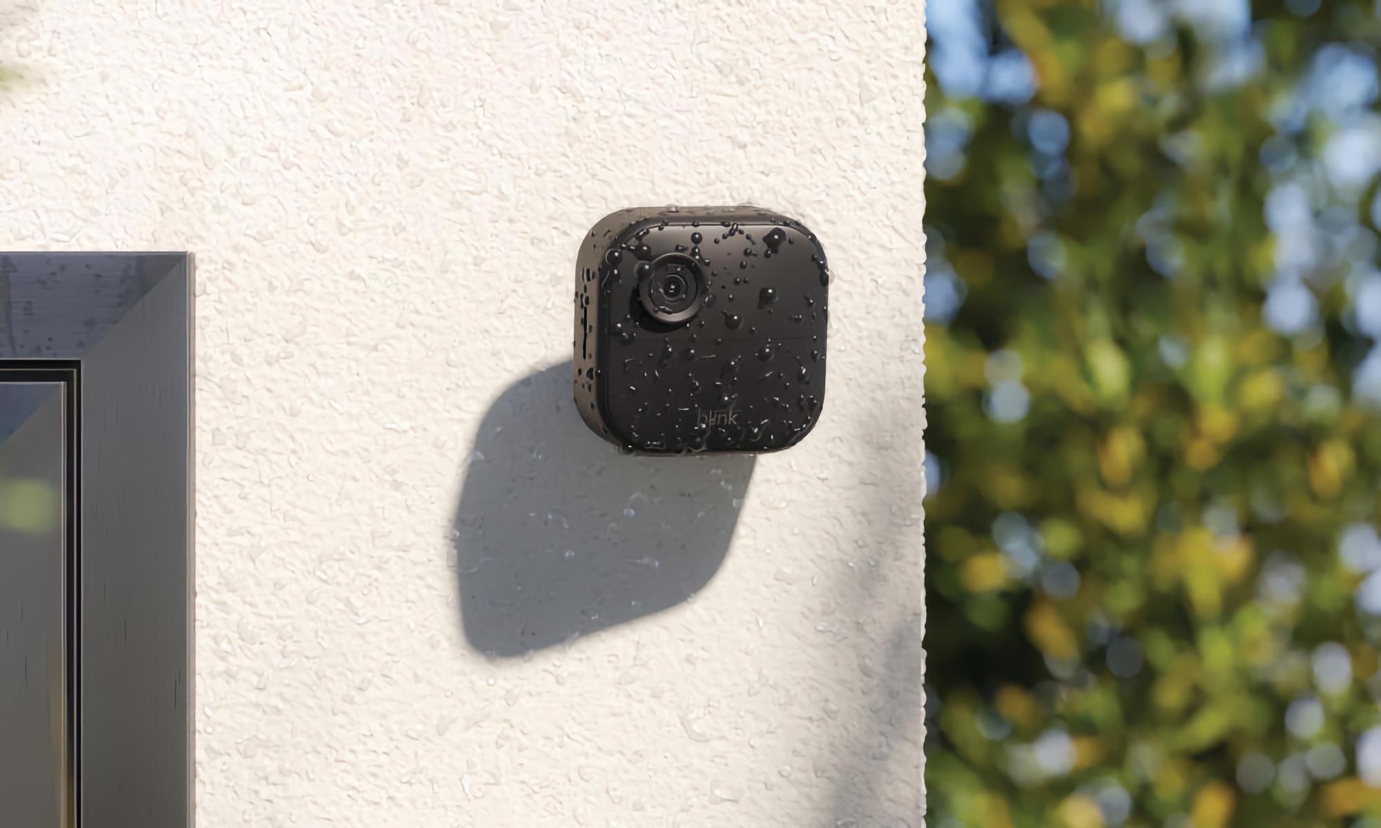 Amazon marketing photo of the Blink Outdoor 4 camera. The black camera has a flat face and rounded corners. It's mounted on a light-colored stucco (outdoor) wall with blurred trees in the background.