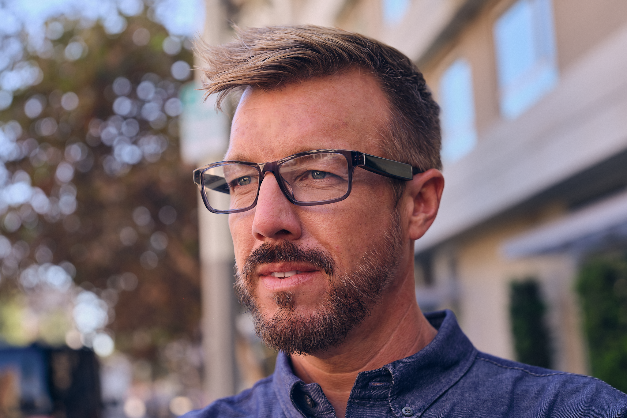Amazon product lifestyle photography of a man wearing Amazon Echo Frames. The black frames look mostly like regular prescription glasses but have very thick arms. He is middle-aged and has a beard and stylish hair. Outdoors with a blurred background.