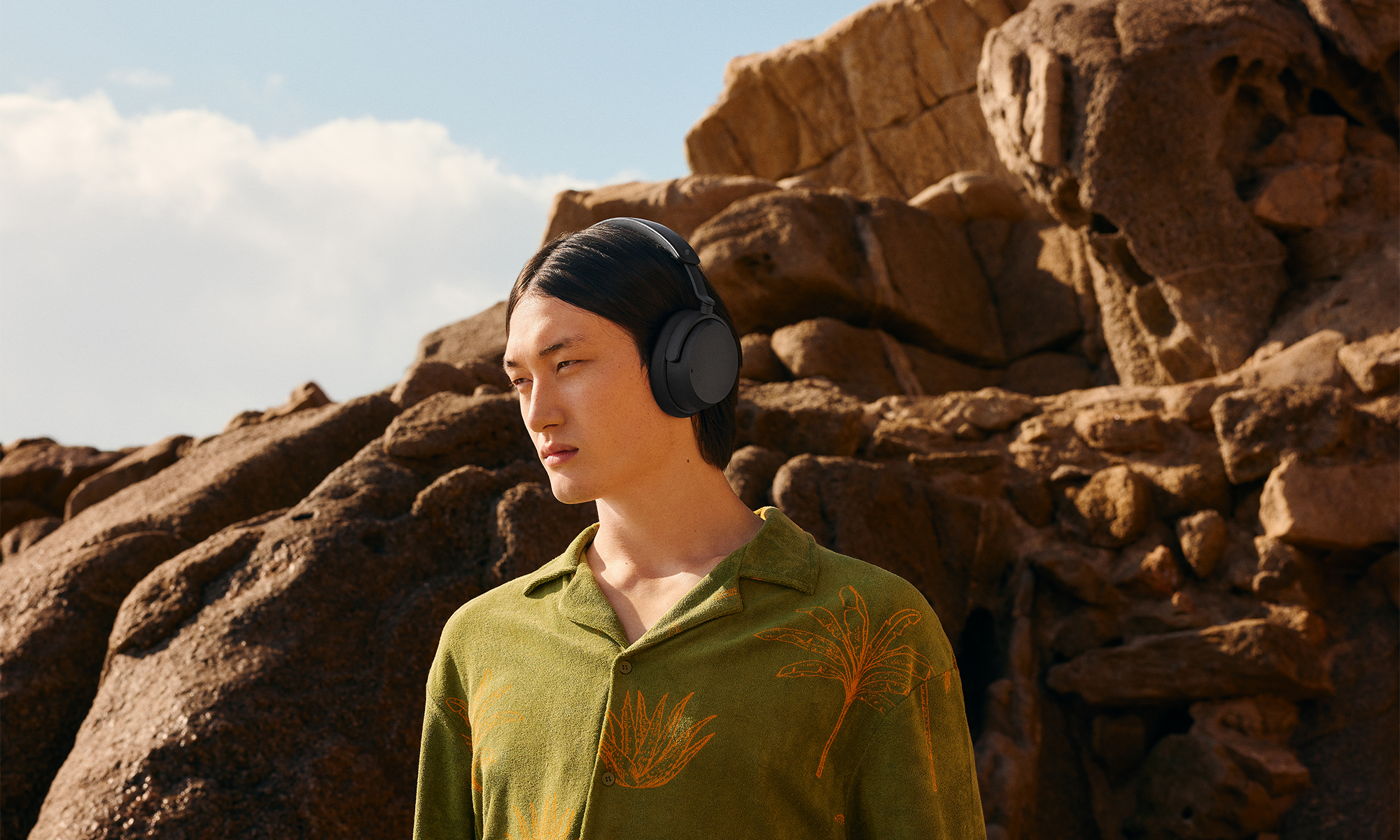 Lifestyle marketing photo for the Sennheiser Accentum Wireless headphones. A young man stands along a rocky terrain: jutting rocks behind him. He wears a green button-down shirt with gold / orange floral patterns.
