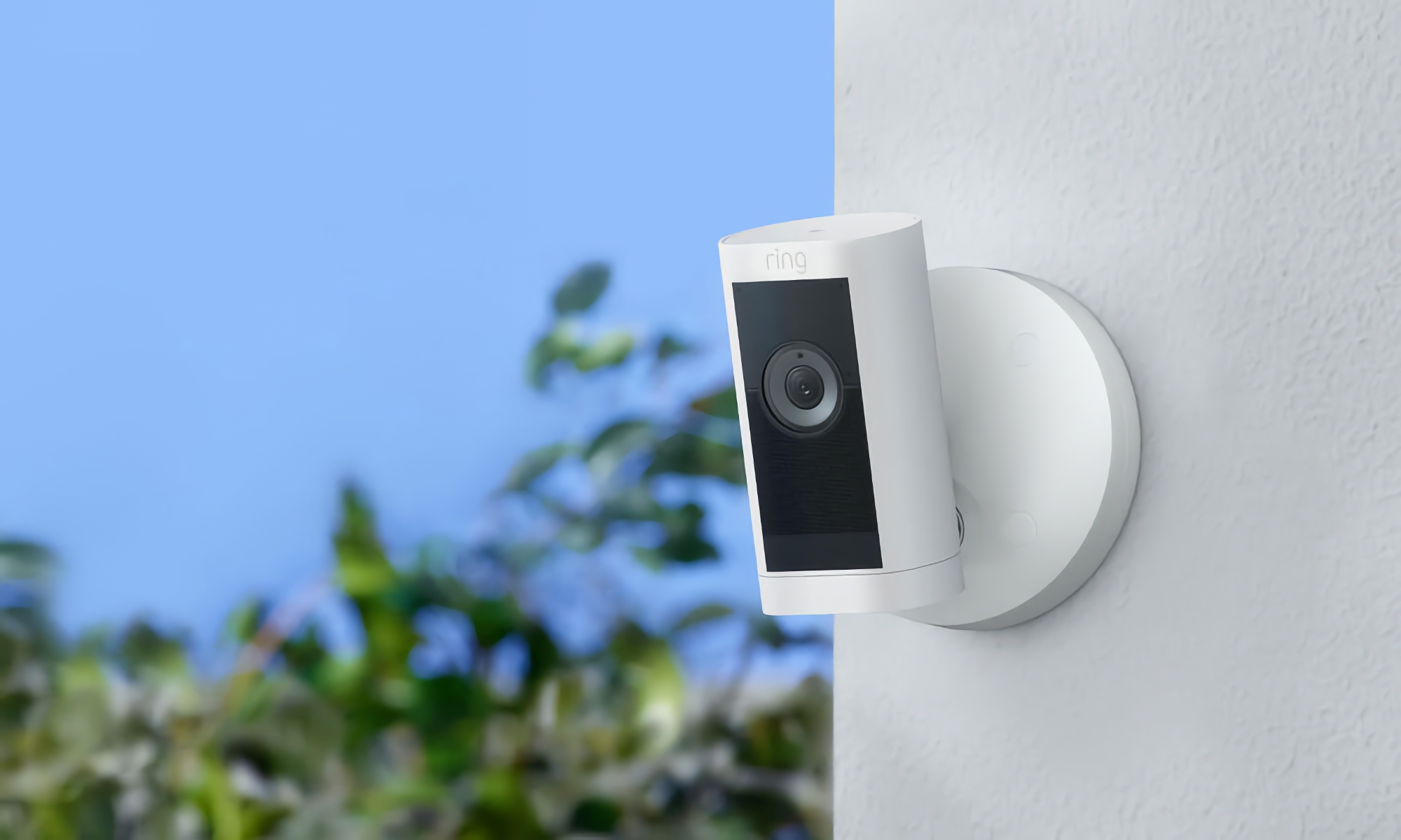 Amazon product photo of the Ring Stick Up Cam Pro. The security camera has a white body and black area near its lens. It's mounted on an outdoor stucco wall with blue sky and green trees visible on the blurred background.